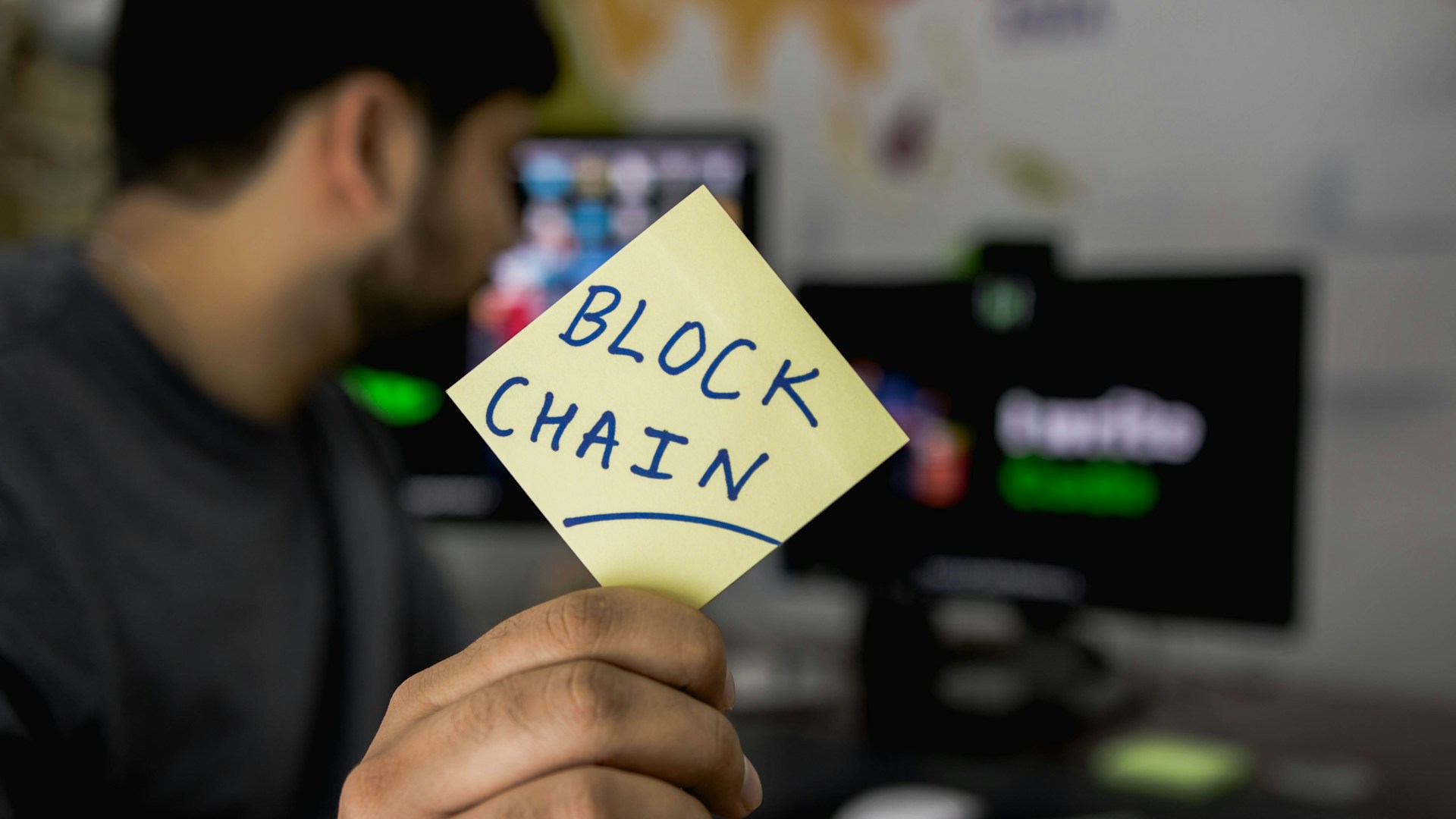 Guy at Computer Holding-Up Sticky-Note that Says “Block Chain”